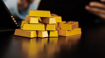 A pile of gold bars on a dark surface with blurred hands and a device in the background.