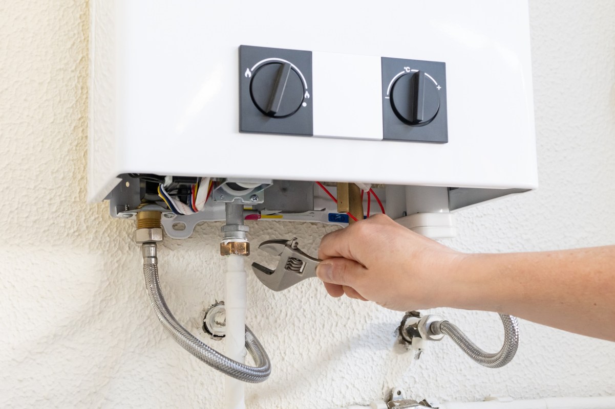 A person uses a wrench to adjust the fittings on a white water heater mounted on a wall.