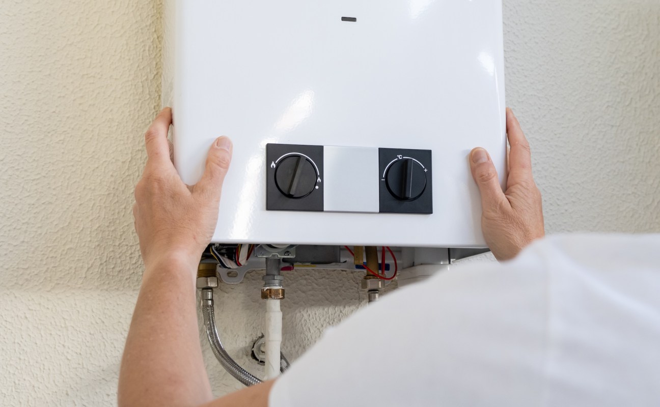 A person adjusts the temperature knobs on a white water heater mounted on a wall.