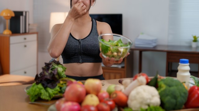 A woman in activewear holds her nose and a bowl of salad while surrounded by various fresh vegetables and fruit on a table.