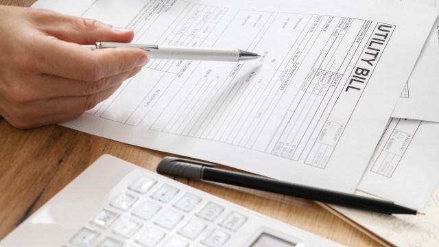 A hand holding a pen points at a utility bill, with a calculator and papers on a wooden desk.