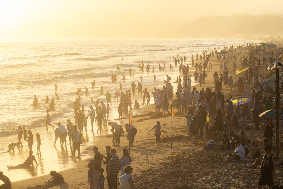 A crowded beach at sunset with people playing in the water and relaxing on the sand.