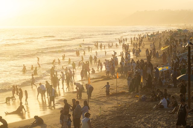 A crowded beach at sunset with people playing in the water and relaxing on the sand.