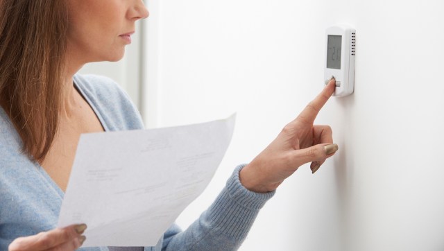 A woman adjusts a thermostat while holding a piece of paper.