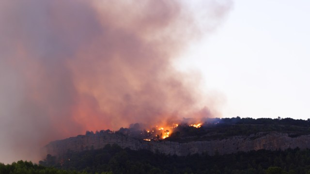 A dense plume of smoke rises from a wildfire on a hillside, with visible flames among the trees.