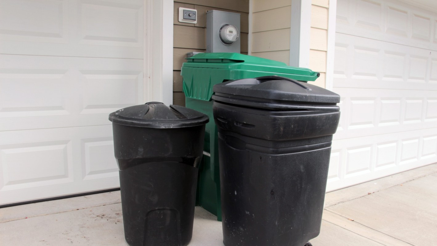 Two trash bins, one black and one green, are placed outside of a garage wall next to a utility meter.