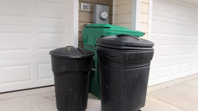 Two trash bins, one black and one green, are placed outside of a garage wall next to a utility meter.