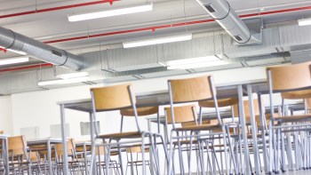 A modern, empty classroom with metal chairs arranged around tables and exposed ductwork on the ceiling.