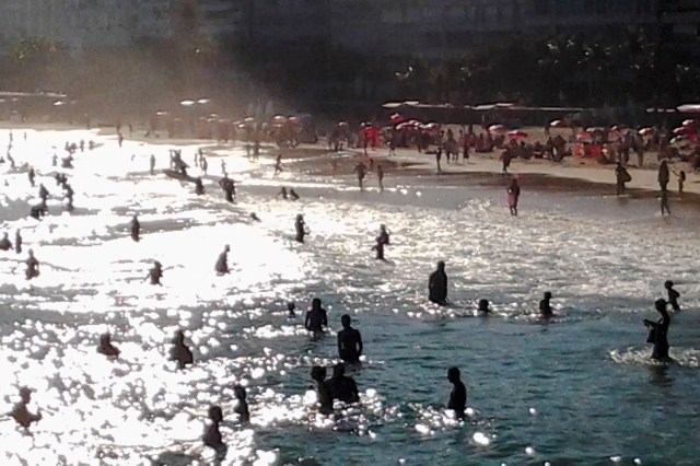 People swimming and playing in the ocean under sunlight, with a beach and umbrellas visible in the background.