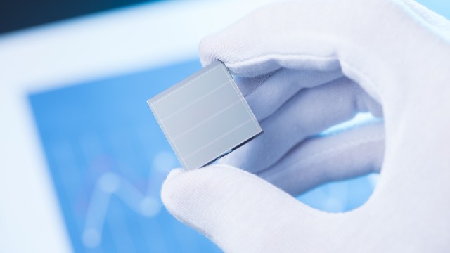 A person in a white glove holds a small, square solar cell against a blurred background.