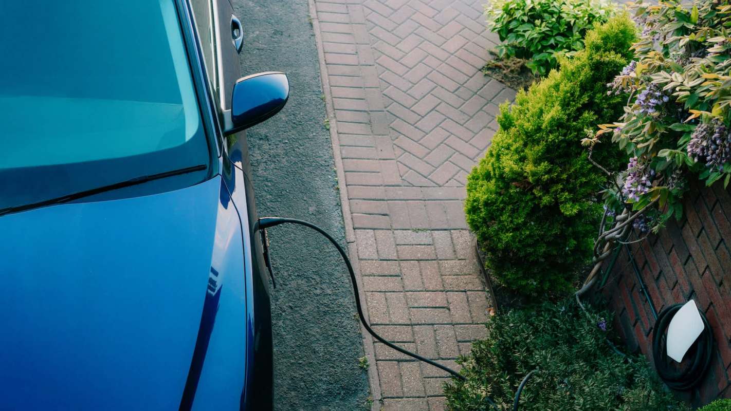 A blue electric car is charging near a neatly paved walkway and greenery.