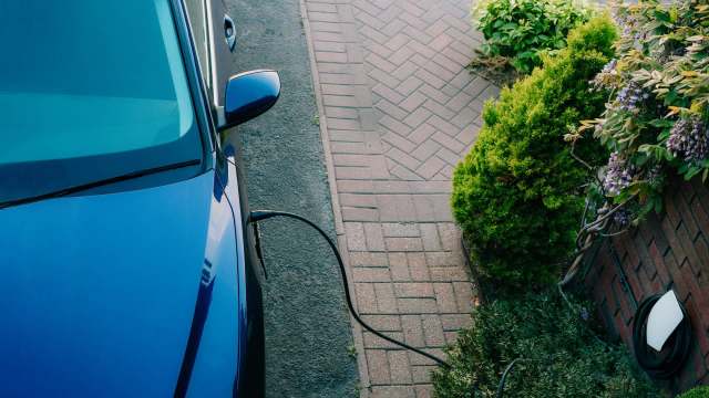 A blue electric car is charging near a neatly paved walkway and greenery.