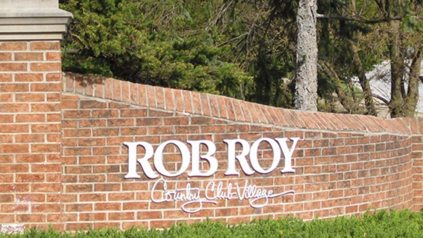 A brick sign reading "Rob Roy Country Club Village" with green hedges and trees in the background.
