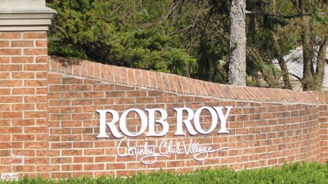 A brick sign reading "Rob Roy Country Club Village" with green hedges and trees in the background.