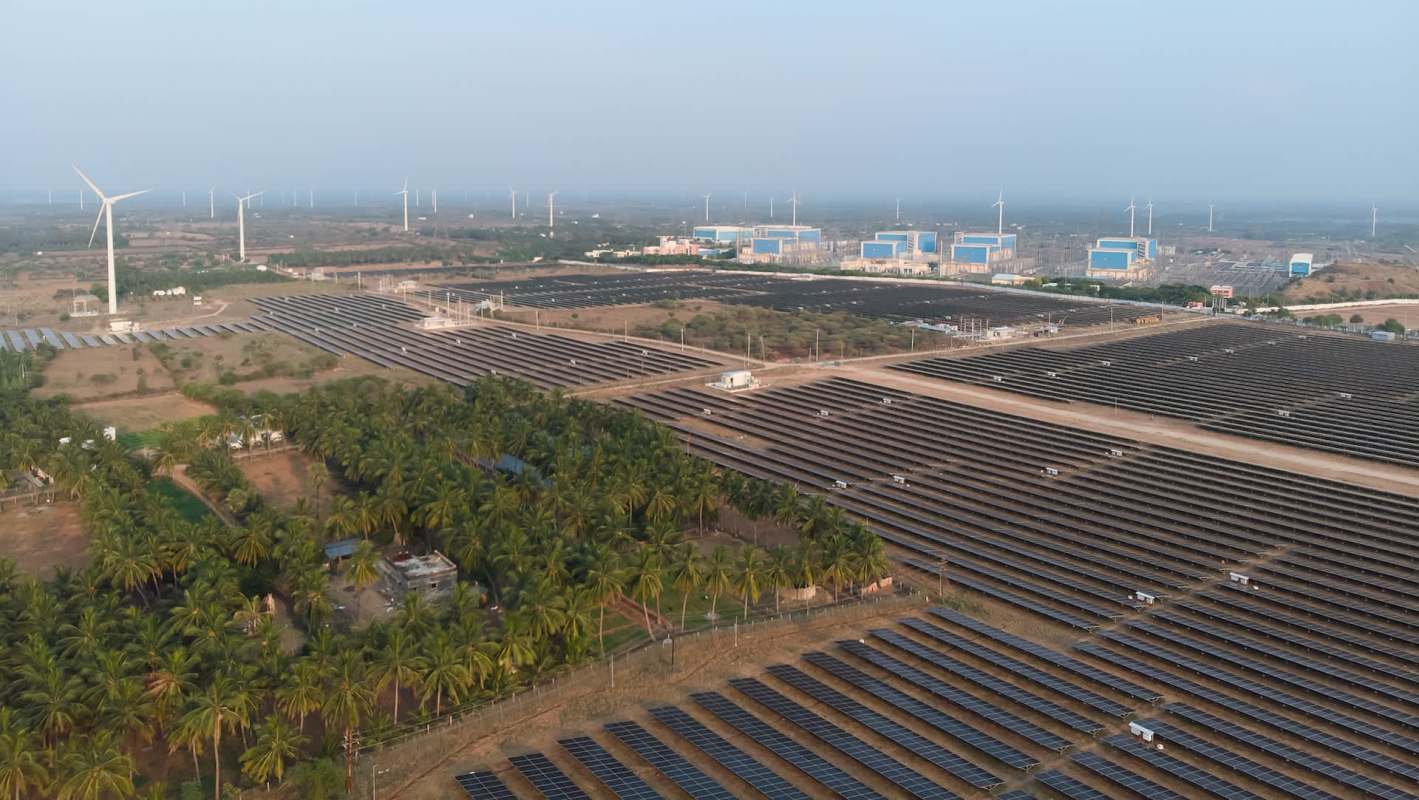 Aerial view of a large solar farm with wind turbines in the background and green fields nearby.