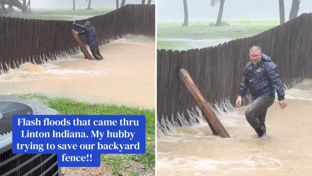 A man in a raincoat struggles against rising floodwaters near a wooden fence during heavy rain.