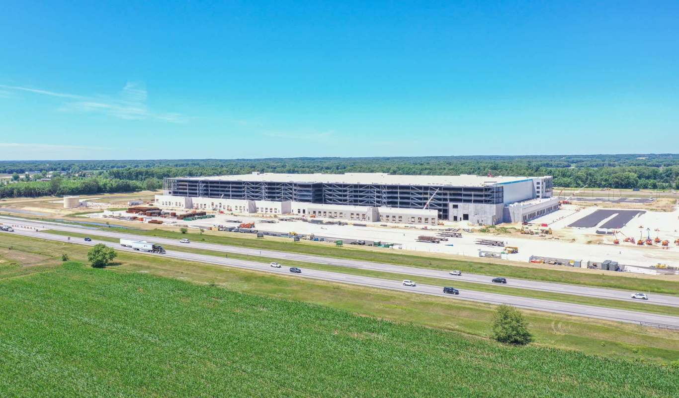 A large industrial building under construction next to a highway, surrounded by farmland and trees.
