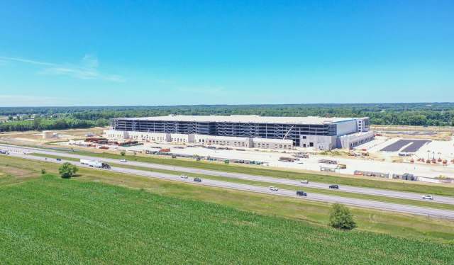 A large industrial building under construction next to a highway, surrounded by farmland and trees.