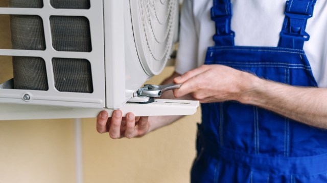 A technician in blue overalls repairs an outdoor air conditioning unit using a wrench.