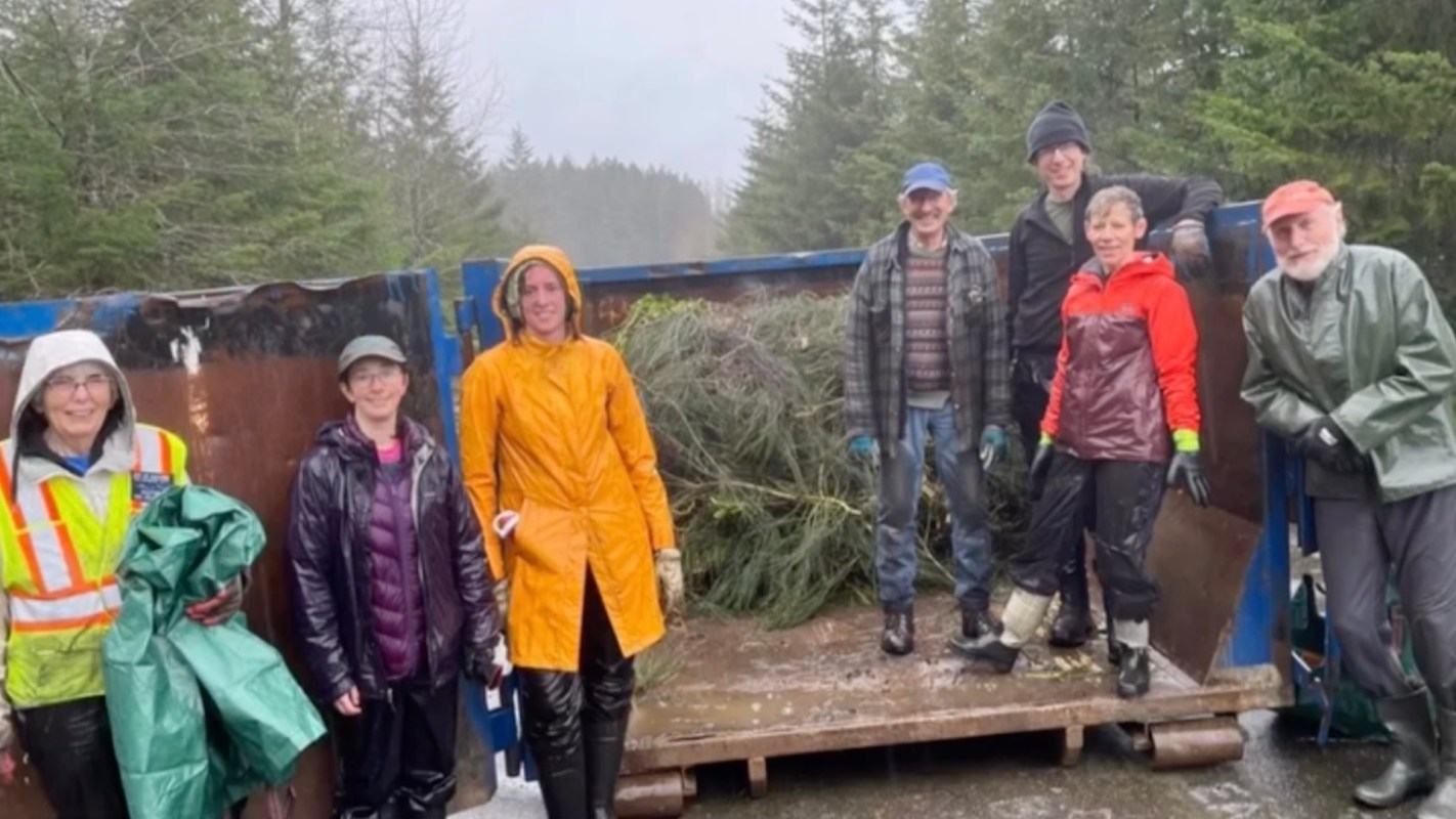 A group of people stands on a vehicle loading area with trees and greenery in the background, all wearing rain gear.