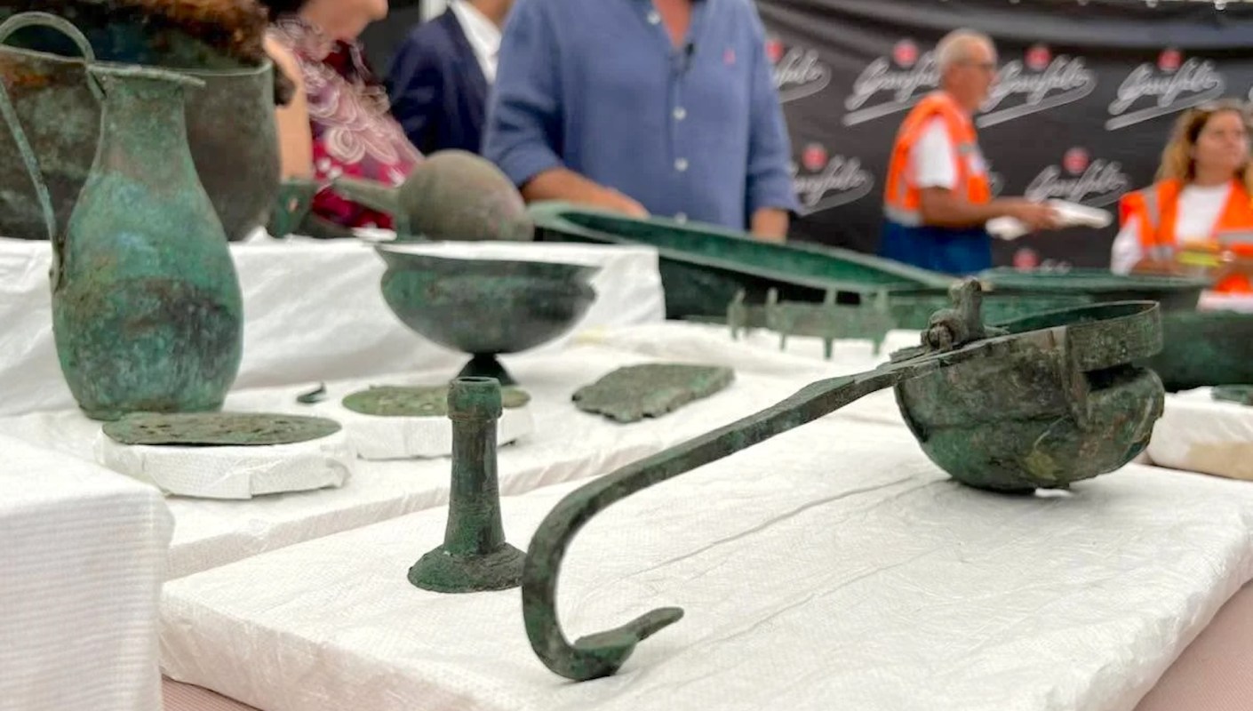 A display of ancient bronze artifacts on a white table, with people in the background.