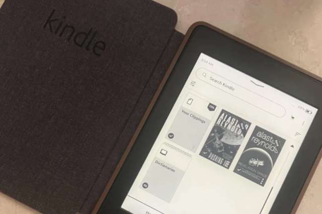 A Kindle e-reader displaying a list of e-books and an open cover on a textured surface.