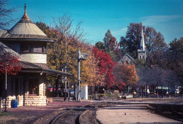 A quaint train station surrounded by vibrant autumn trees and a distant church steeple.