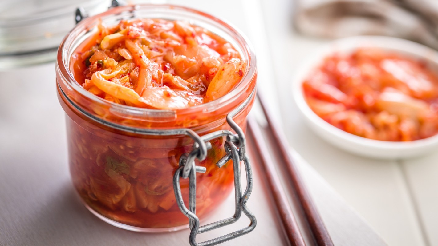 A glass jar filled with vibrant kimchi sits on a table with wooden chopsticks nearby.