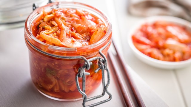A glass jar filled with vibrant kimchi sits on a table with wooden chopsticks nearby.