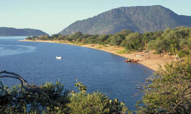 A tranquil shoreline with a boat on the water, surrounded by lush greenery and distant hills.