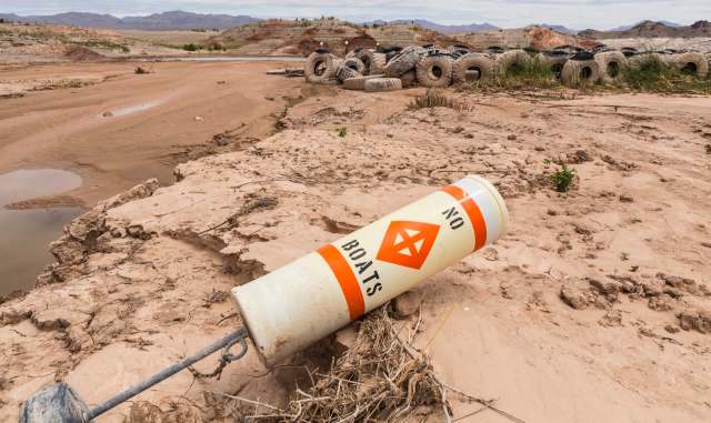 A sandy shore with a "No Boats" buoy lying on the ground, surrounded by dry landscape and scattered tires.