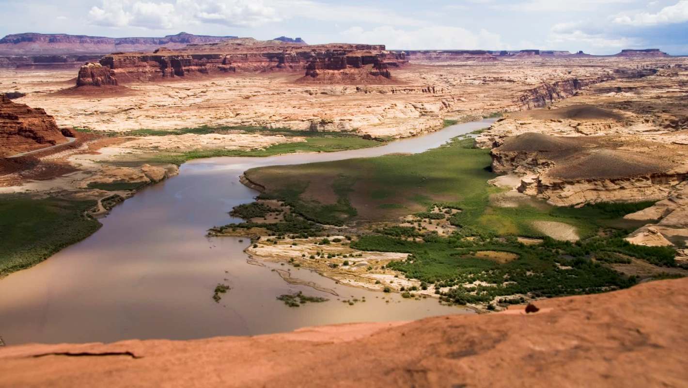 A panoramic view of a winding river surrounded by red rock formations and green vegetation in a desert landscape.