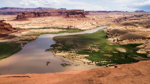 A panoramic view of a winding river surrounded by red rock formations and green vegetation in a desert landscape.