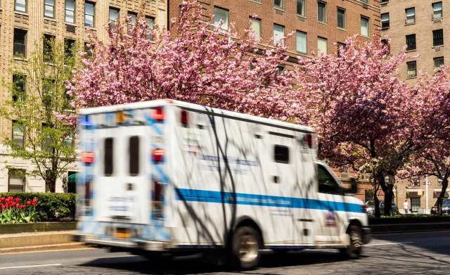 An ambulance drives past blooming cherry blossom trees and urban buildings on a sunny day.