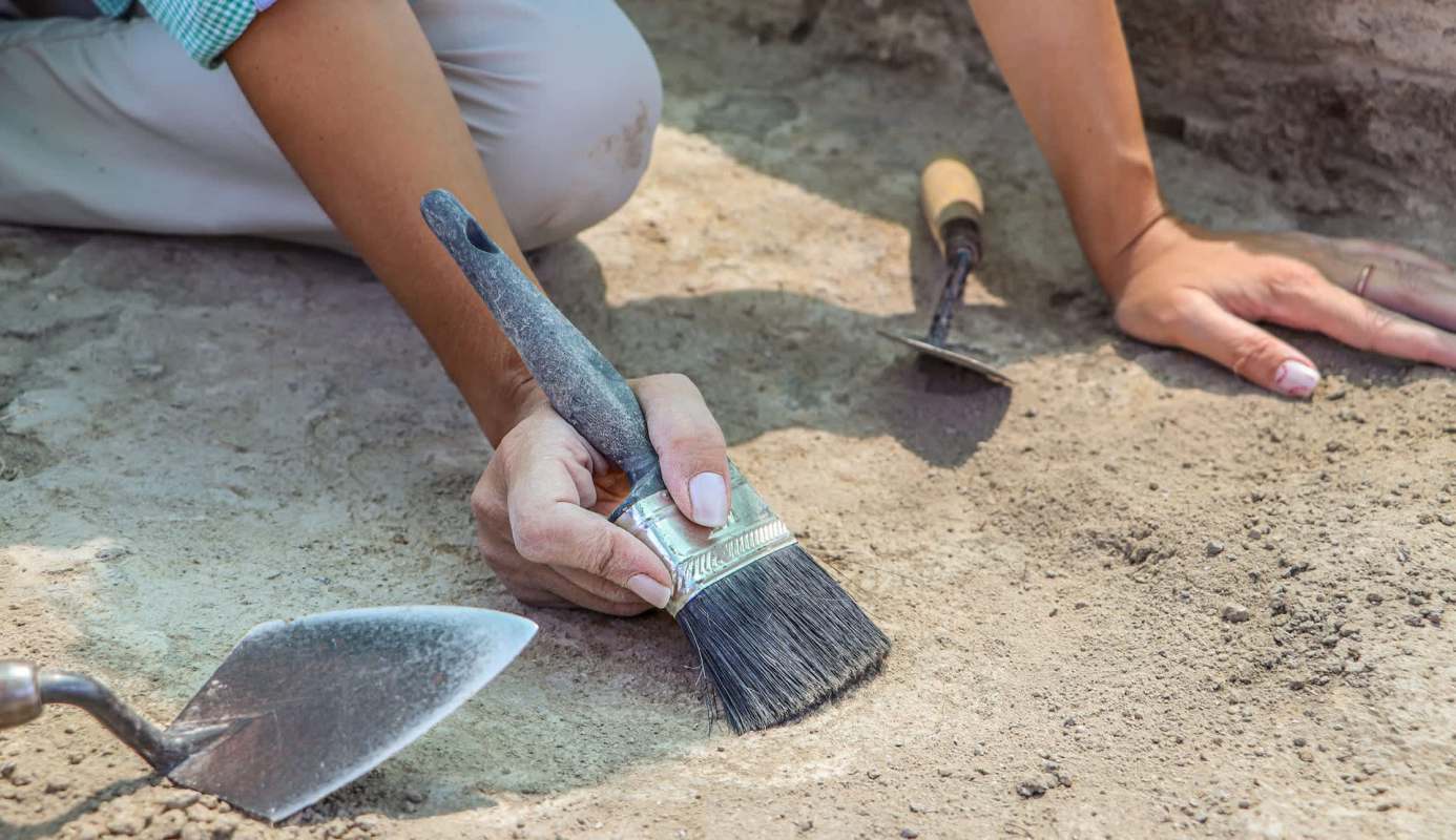 A person using a brush to carefully uncover artifacts in a dusty excavation site, surrounded by tools.
