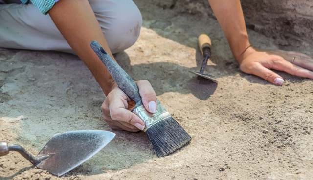 A person using a brush to carefully uncover artifacts in a dusty excavation site, surrounded by tools.