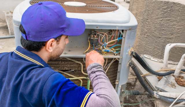 A technician repairs an air conditioning unit, examining wiring and components outside a building.