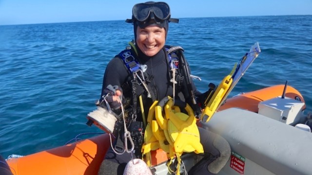A smiling diver wearing a wetsuit holds diving equipment on the edge of a boat in clear blue water.