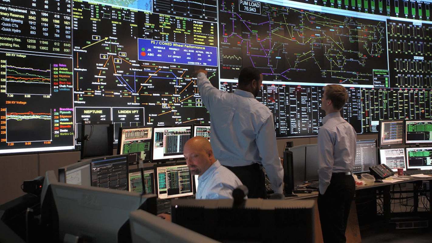 Three individuals analyze data on multiple large screens in a control room filled with computer monitors.