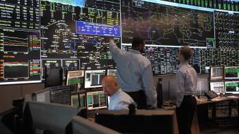 Three individuals analyze data on multiple large screens in a control room filled with computer monitors.