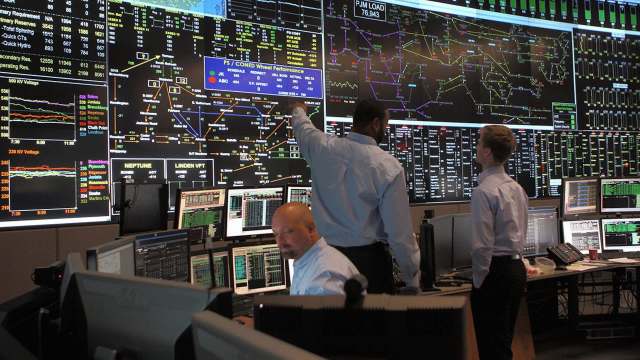Three individuals analyze data on multiple large screens in a control room filled with computer monitors.