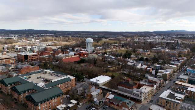 An aerial view of a small town featuring buildings, a water tower, and distant hills under a cloudy sky.