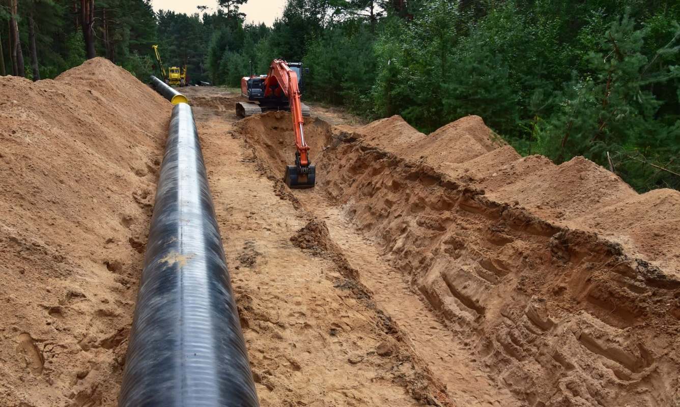 Excavation site with a large black gas pipeline laid in a trench, alongside heavy machinery and piles of dirt.