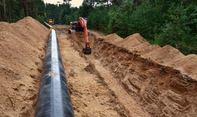 Excavation site with a large black gas pipeline laid in a trench, alongside heavy machinery and piles of dirt.
