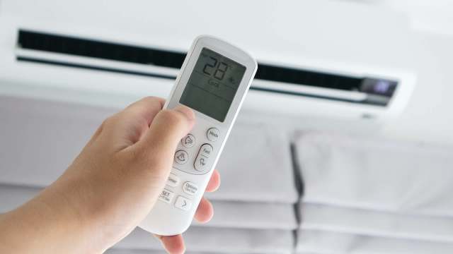 A person holds a remote control displaying 28°C aimed at an air conditioner unit.