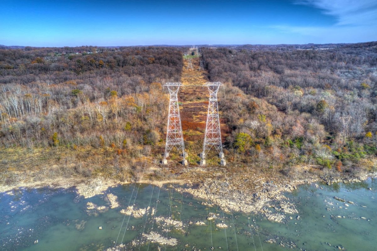 Aerial view of power lines towering over a forested area, with a river and rocky shoreline below.