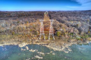 Aerial view of power lines towering over a forested area, with a river and rocky shoreline below.