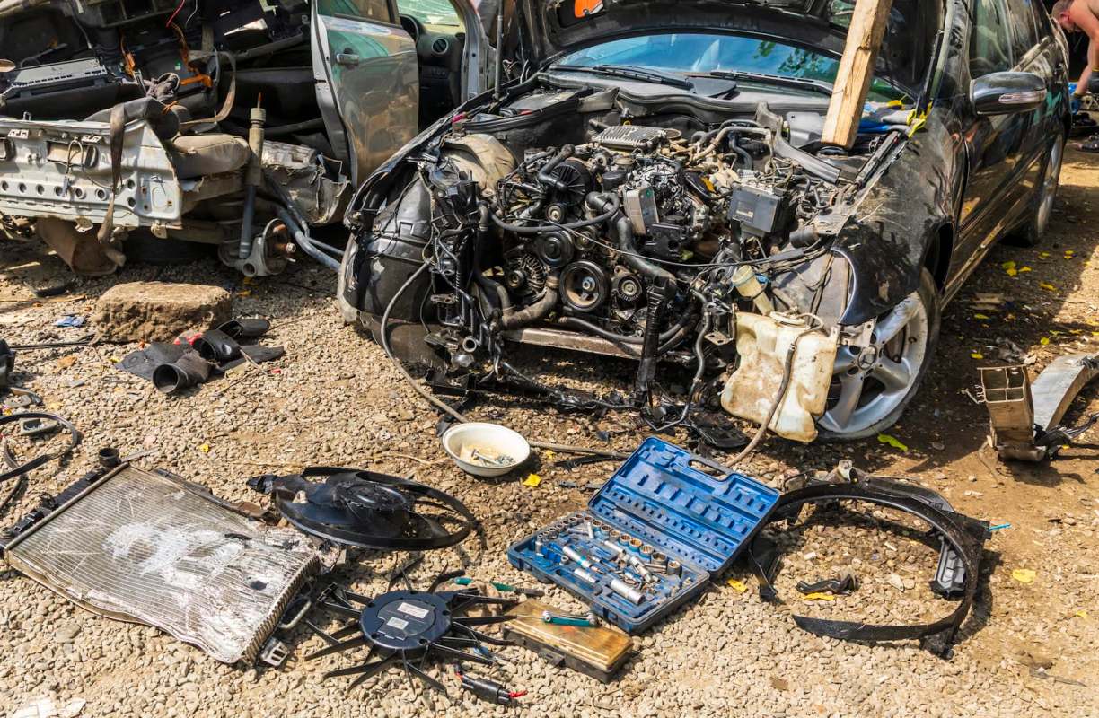A dismantled car in a junkyard with engine parts and tools strewn across a gravel surface.