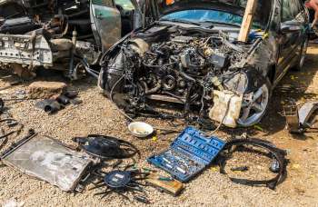 A dismantled car in a junkyard with engine parts and tools strewn across a gravel surface.