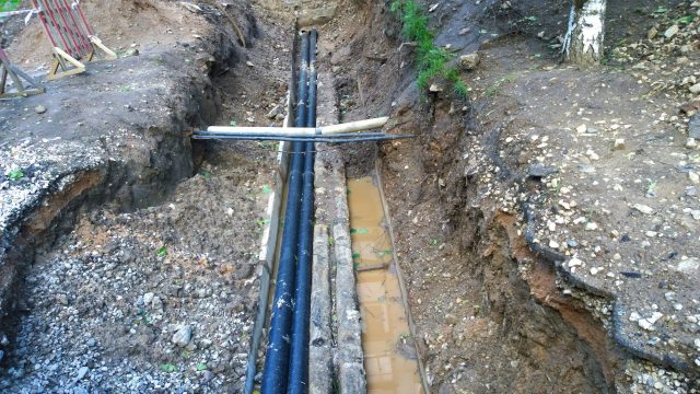 A deep trench with black pipes and muddy water, surrounded by rough soil and scattered stones.